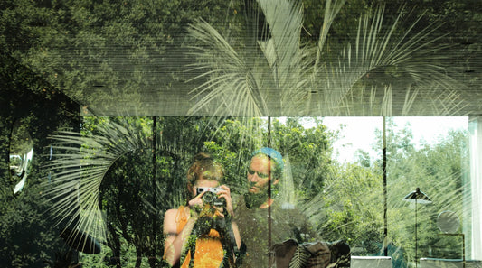 A double exposure film photo of a couple taking a selfie in front of reflective glass with plants and showing the interior of a modern home